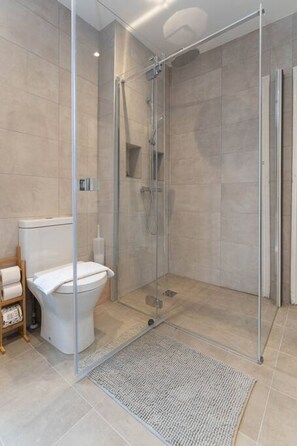 A modern bathroom with a glass-enclosed shower, a toilet, and a small shelf with toiletries.