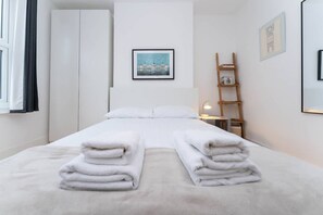 Un letto rifatto con asciugamani piegati, un comodino con una lampada, un quadro appeso alla parete e una scala appoggiata al muro.