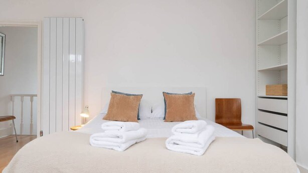 Una cama bien hecha con toallas blancas dobladas, una silla de madera y un armario blanco con estantes.
