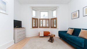 Una sala de estar con televisor, un sofá azul, una mesa de centro de madera y una ventana con contraventanas de madera.