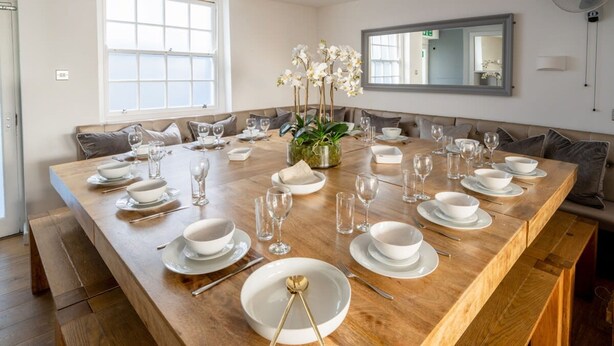 A large wooden dining table set for six with white plates, bowls, and glasses, surrounded by cushioned seating. - Host Stay The Charles Street Retreat (Brighton)