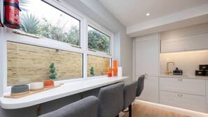 Une cuisine moderne avec des meubles blancs, un coin bar doté de tabourets gris et une fenêtre donnant sur une clôture en bois et de la verdure.