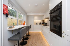 A modern kitchen with white cabinetry, a double oven, and a bar area with stools. - Host Stay Brighton Laines House (Brighton)