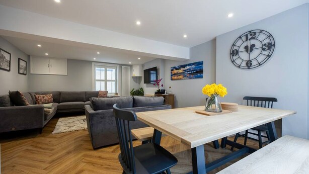 Un salon moderne comprenant un ensemble de canapés, une table à manger avec des chaises, une horloge au mur et un téléviseur fixé au-dessus du canapé.