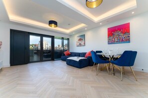 A modern living room with a sofa, dining table, and chairs. There are two paintings on the wall, a sliding glass door, and a view of the city. - Host Stay Valley Gardens Apartment (Brighton)