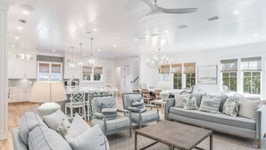 A spacious living room with a grey sofa, matching armchairs, and a coffee table. A dining area with a chandelier and a kitchen with white cabinets and a large window. - Mischievous Mermaid|Easy Beach Access|Private Pool|Game Room|Golf Cart (Seagrove Beach)