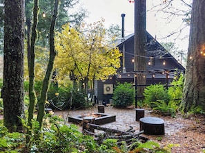 A rustic cabin with a stone fireplace, surrounded by a forested area with string lights.