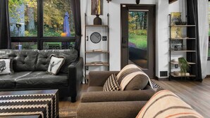 Een woonkamer met een leren bank, een bruine fauteuil en een vloerkleed met een patroon. Er is een boekenkast met decoratieve objecten en uitzicht op een tuin door een glazen deur.