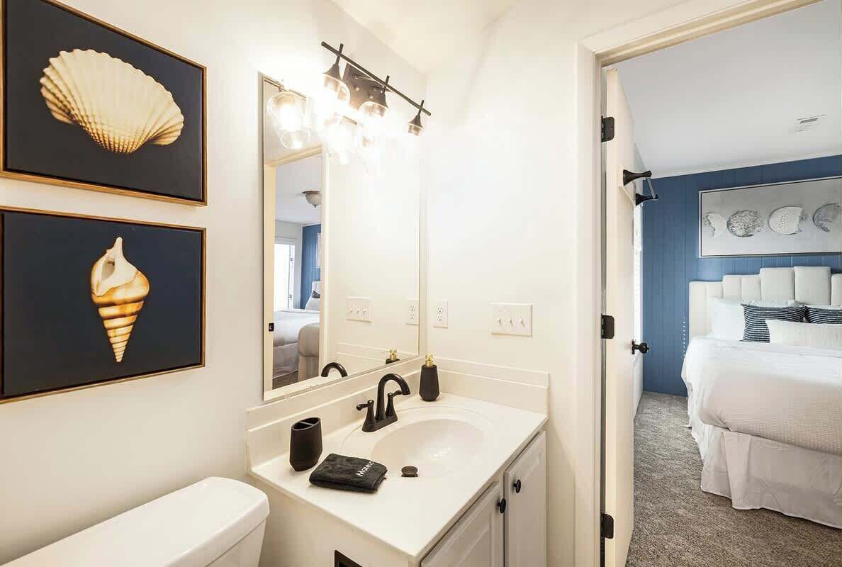 A hotel bathroom with a white sink, black faucet, and a mirror above it. There are framed pictures on the wall and a bed visible in the adjacent room.