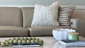Ett vardagsrum med en beige soffa, mönstrade kuddar, ett soffbord med böcker och en grön kruka, samt konstverk på väggen ovanför soffan.