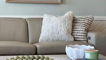 A living room with a beige sofa, patterned cushions, a coffee table with books and a green container, and wall art above the sofa.