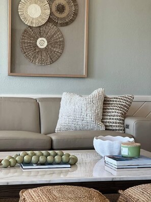 A living room with a beige sofa, two patterned throw pillows, a coffee table with a book and a green container, and decorative wall art. - Oceanfront Free EV charger & GolfCart, Pool (Ocean Isle Beach)