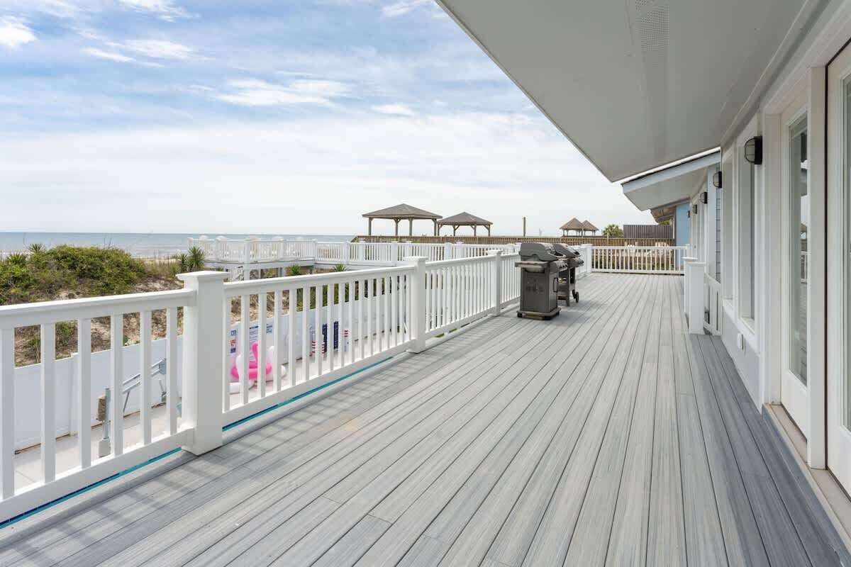 A wooden deck with a white railing, overlooking a beach with umbrellas and a grill.