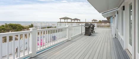 A wooden deck with a white railing, overlooking a beach with umbrellas and a grill.