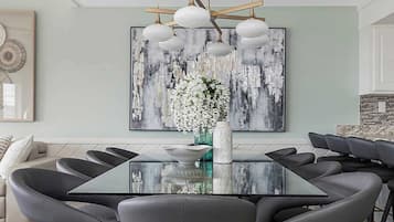 A modern dining area with a glass table surrounded by chairs, a large abstract painting on the wall, and a chandelier above.