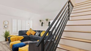 A living room with a dark grey sectional sofa, yellow and blue cushions, a wooden staircase, and a potted plant.