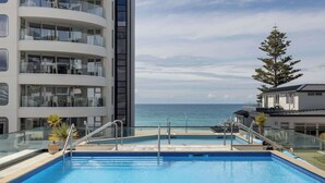 Una piscina con barandilla de cristal, rodeada por un edificio moderno con balcones y vistas al mar.