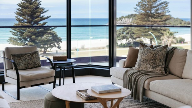 Un salón moderno con un ventanal grande que da a una playa y árboles. Hay un sofá, dos sillones, una mesa de centro y una mesita con un libro.