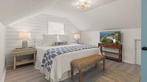 A bedroom with a sloped ceiling, a bed with a white and blue patterned bedspread, a television on a wooden stand, and a wicker bench. - Toddy Cottage located on East Beach, sleeps 10 (St Simons Island)