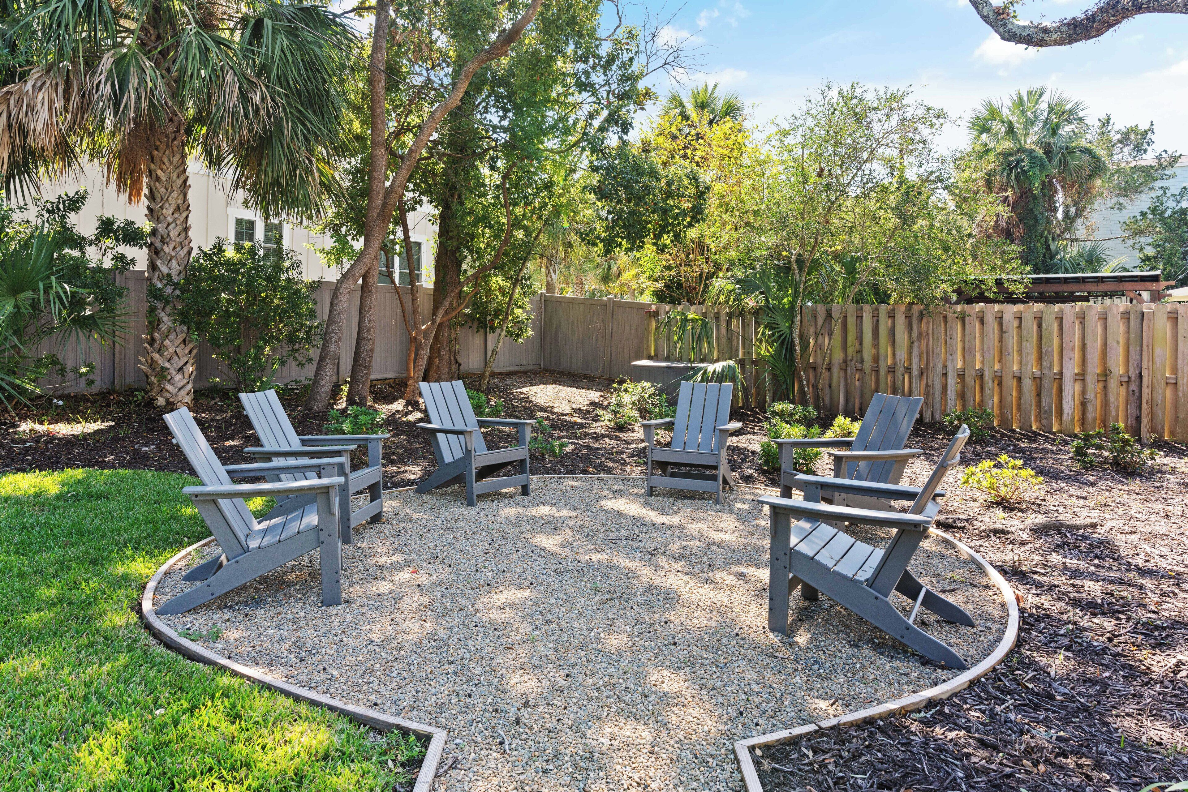A patio with four Adirondack chairs arranged in a semi-circle, surrounded by gravel and grass, with a wooden fence and trees in the background.