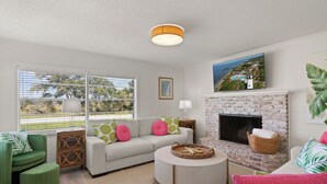A modern living room with a fireplace, a sofa set, a round coffee table, and a view of the outdoors through large windows. - Toddy Cottage located on East Beach, sleeps 10 (St Simons Island)