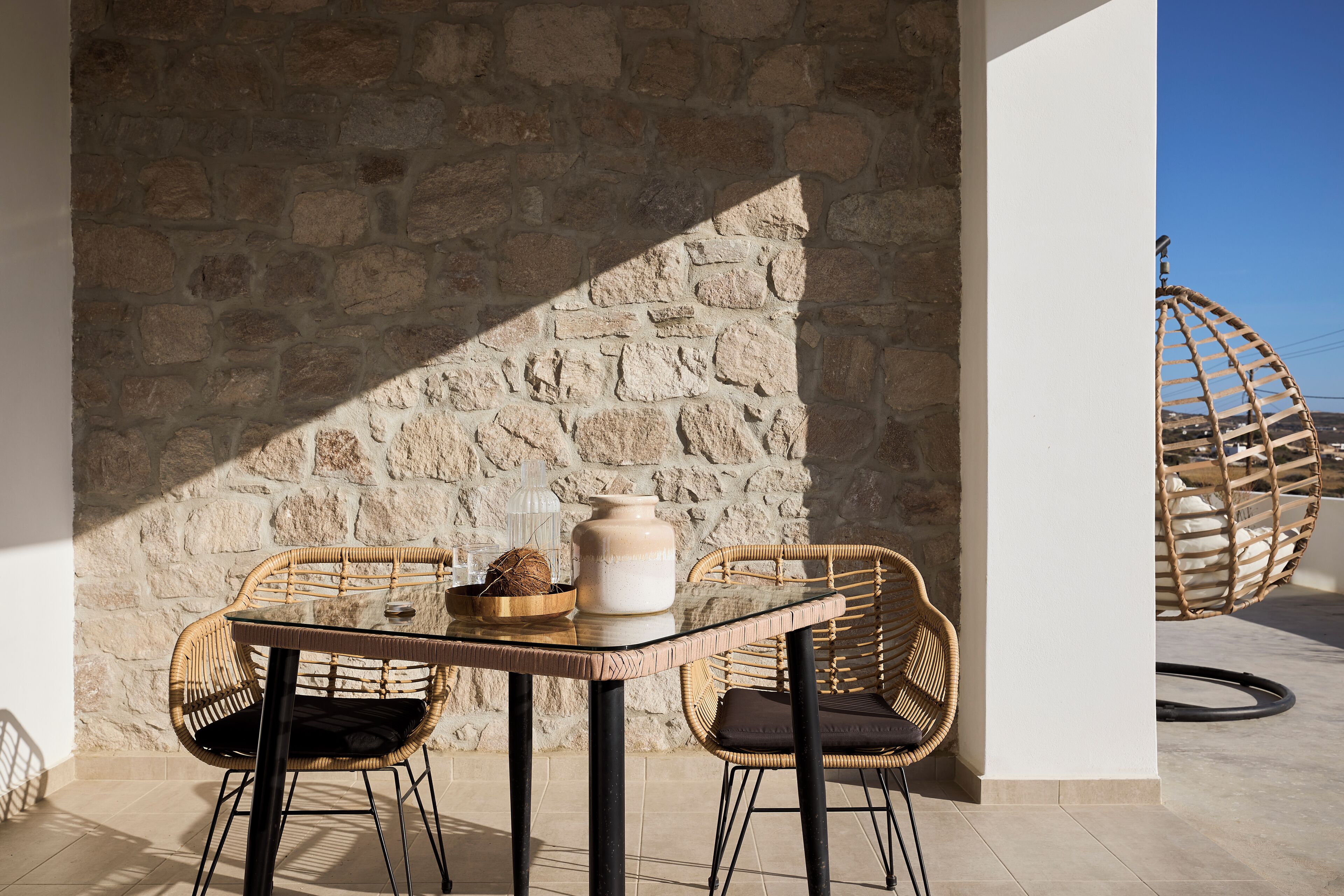 Un muro de piedra, sillas de mimbre, una mesa de vidrio y un sillón colgante en un espacio exterior iluminado por el sol.
