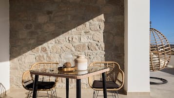 Un muro de piedra, sillas de mimbre, una mesa de vidrio y un sillón colgante en un espacio exterior iluminado por el sol.