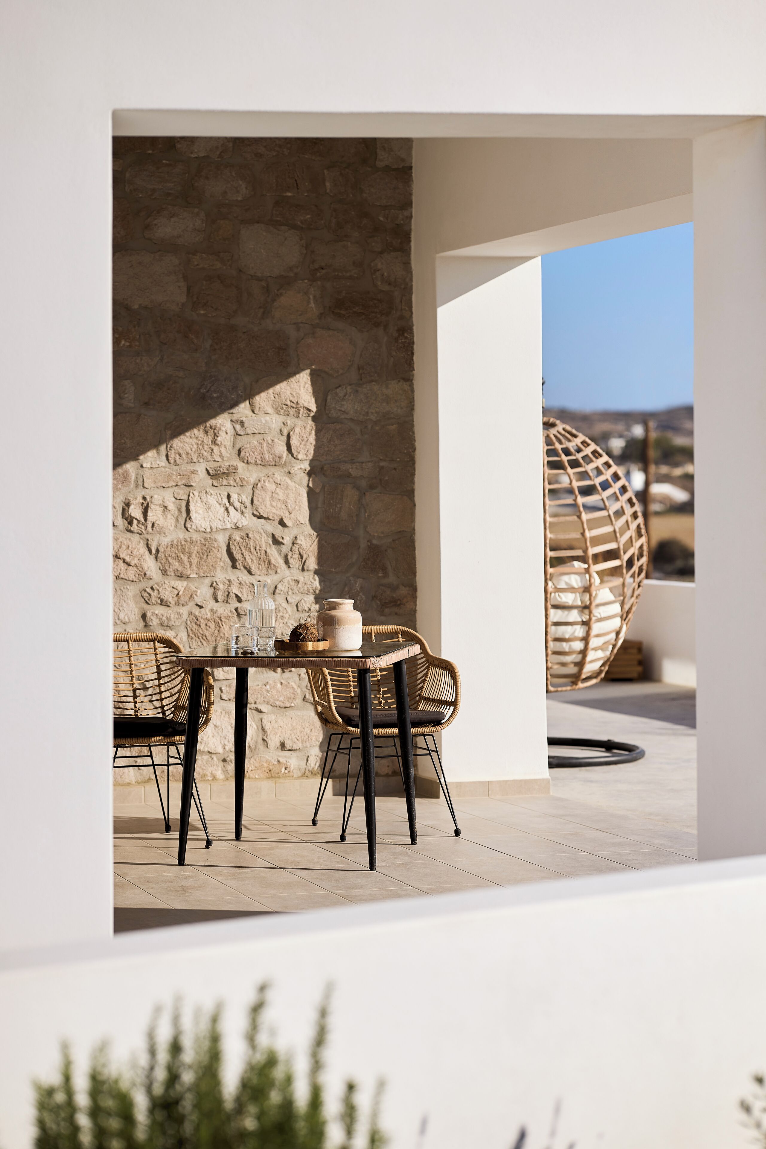 Un espacio al aire libre con pared de piedra, sillas de mimbre y una mesa con un florero y una vela.