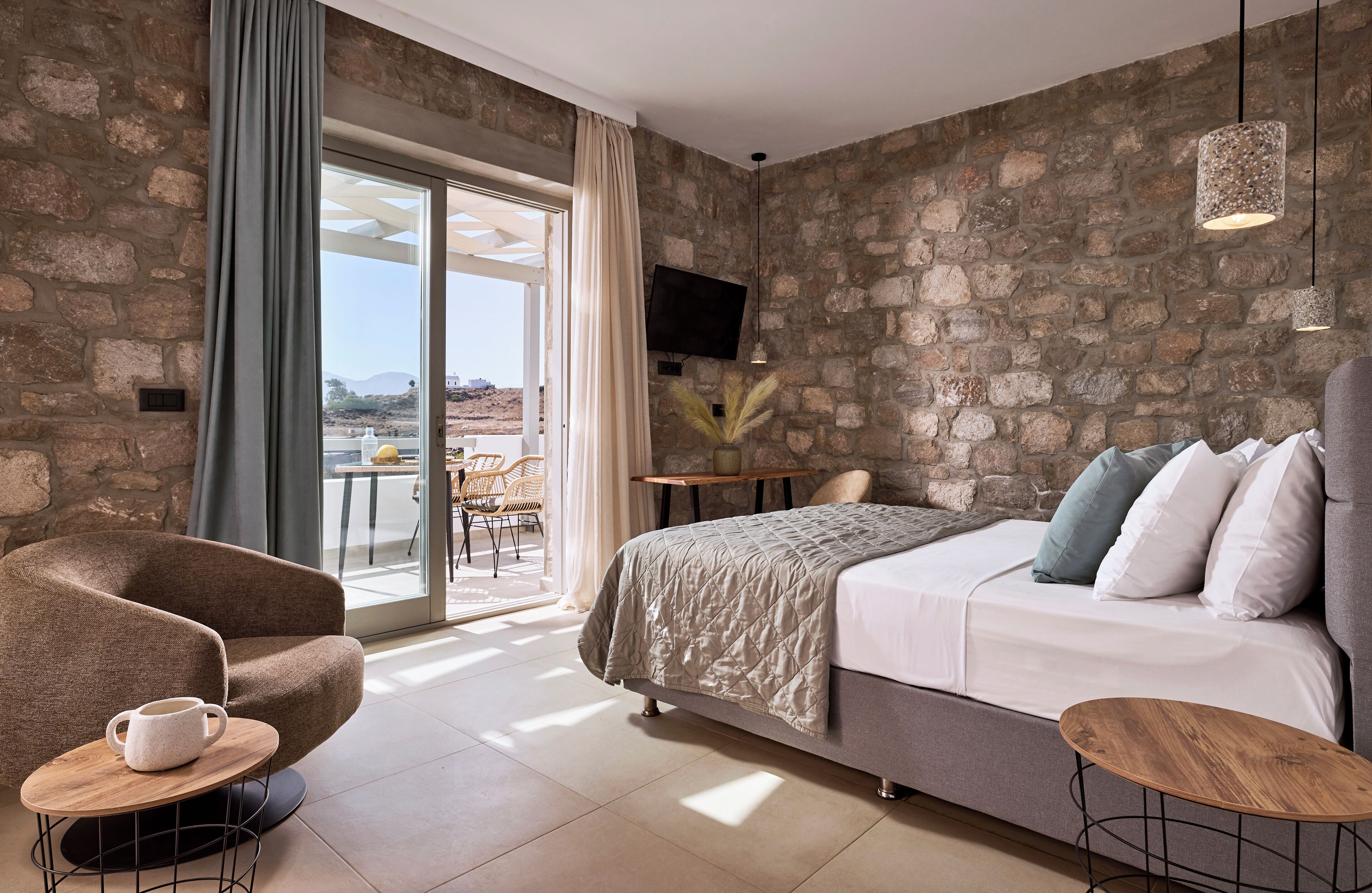 Une chambre moderne avec un mur en pierre, un lit agrémenté de plusieurs coussins, une chaise, une petite table et un téléviseur fixé au mur.