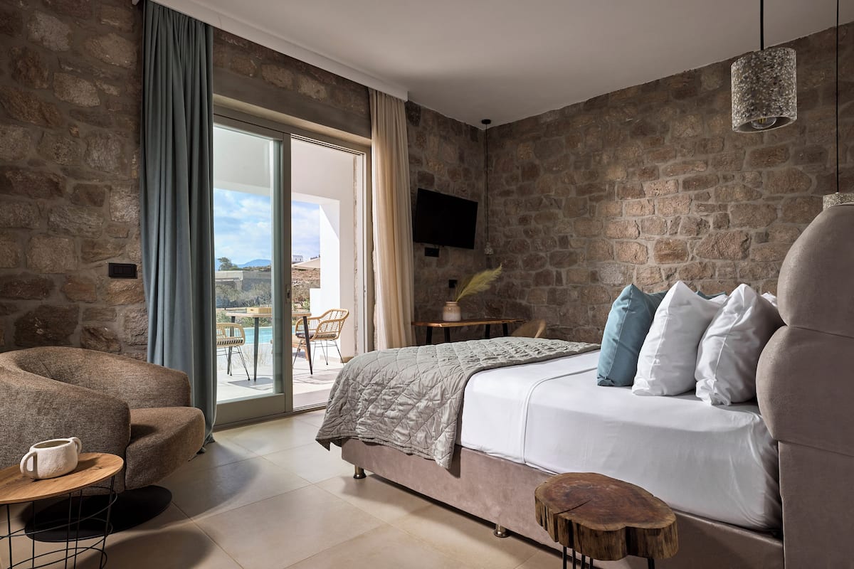 A bedroom with a stone wall, a bed with white and blue pillows, a wooden coffee table, and a TV on the wall.