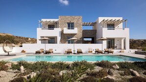 A modern two-story house with a swimming pool, outdoor seating, and a view of the hills. - Kedros Milos Suites (Milos)