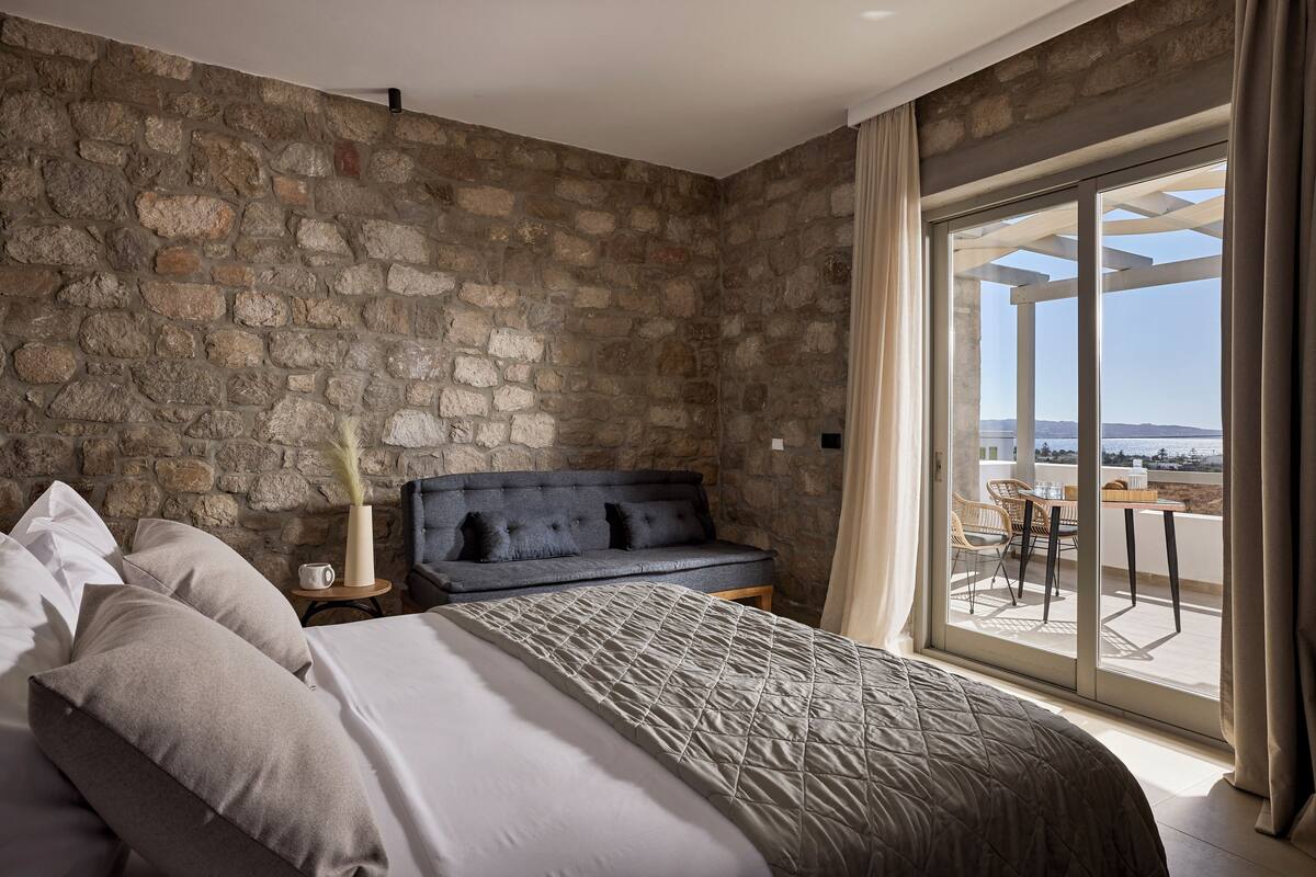 A bedroom with a stone wall, a bed with a quilted bedspread, a dark sofa, and a view of the outdoors through a glass door.