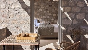 A stone-walled room with a bed, a wicker chair, and a table with a tray of coconuts. - Kedros Milos Suites (Milos)