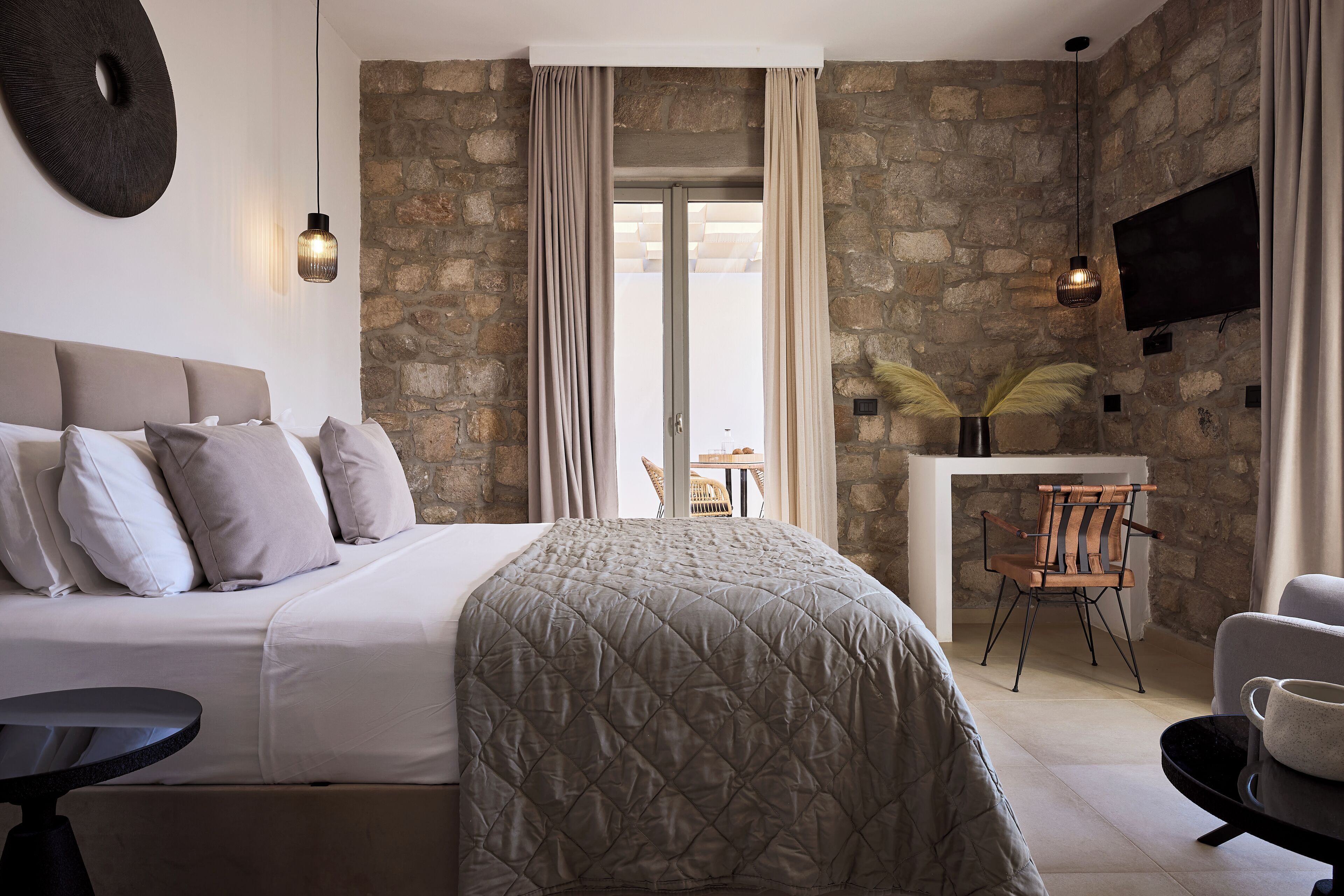 A modern hotel room with a stone wall, a bed with a quilted cover, a desk with a chair, and a TV mounted on the wall.