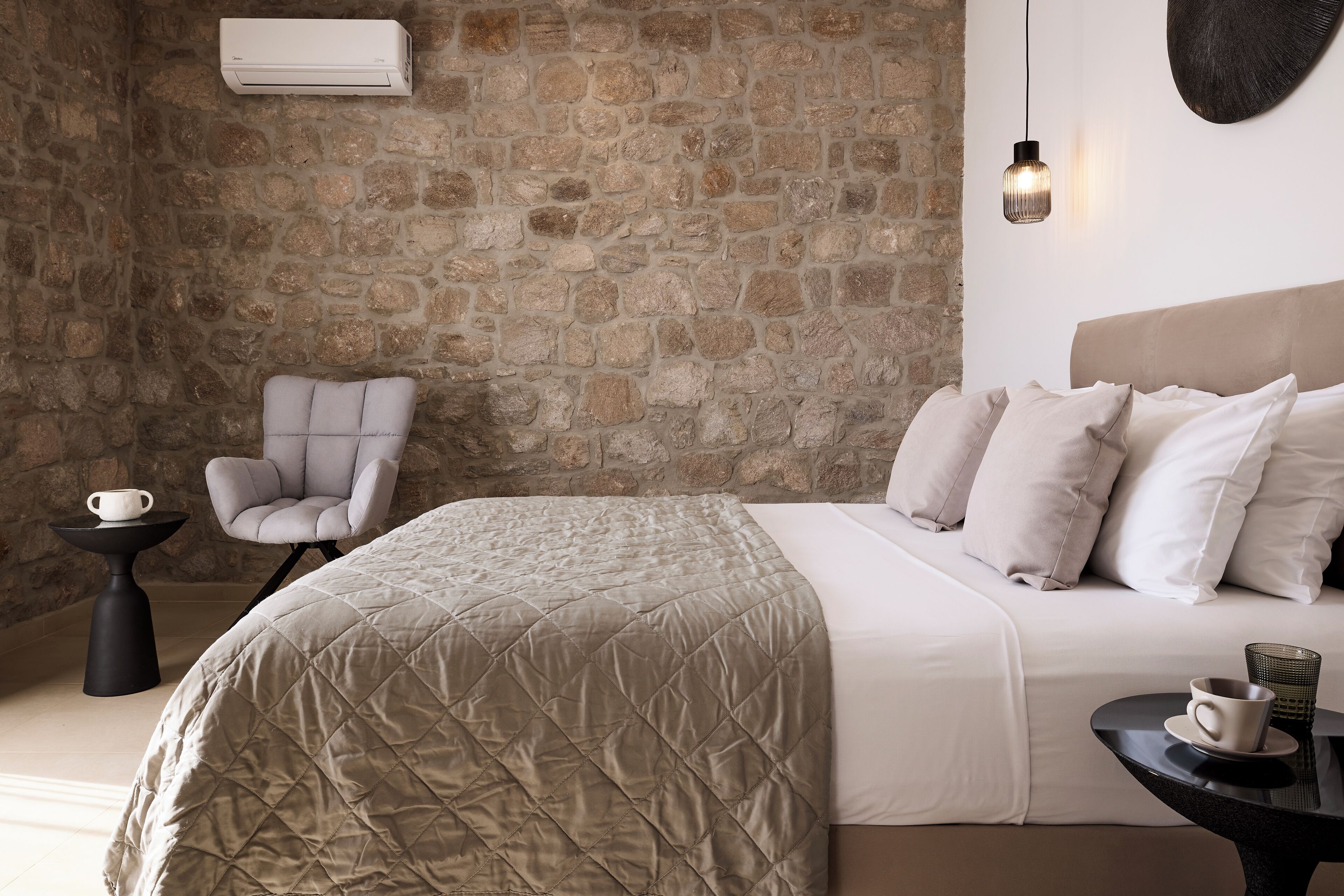 A bedroom with a stone wall, a bed with a quilted cover, a chair, a small table with a cup, and a hanging light fixture.