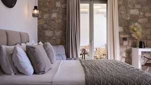 A bedroom with a stone wall, a bed with a quilted blanket, and a view of the outdoors through a glass door. - Kedros Milos Suites (Milos)