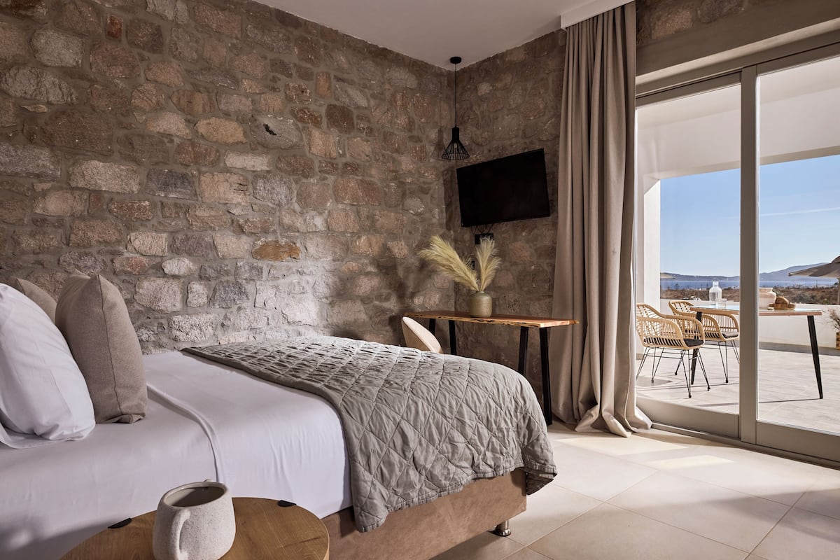 A bedroom with a stone wall, a bed with a quilt, a TV, a table with a vase, and a view of the outdoors.