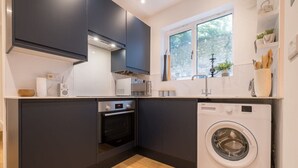 A modern kitchen with dark cabinets, a built-in oven, a washing machine, and a window with a view of greenery. - Host & Stay | Osbourne Rest (Brighton and Hove)