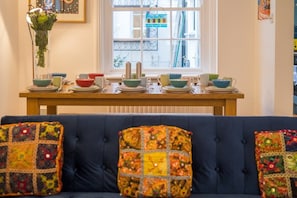 A dining area with a wooden table set for a meal, a blue sofa with colorful cushions, and a window with a view of buildings and a balcony. - Host & Stay | Over Arches (Brighton and Hove)