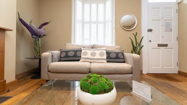Un salon avec un canapé beige, une table basse en verre ornée d’un arrangement de mousse, une fenêtre avec des stores, un miroir rond et un cactus.