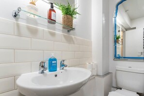 A bathroom with a white sink, two faucets, a glass shelf with toiletries, and a mirror with a blue frame. - Host & Stay | Sea View Marine Parade (Brighton and Hove)