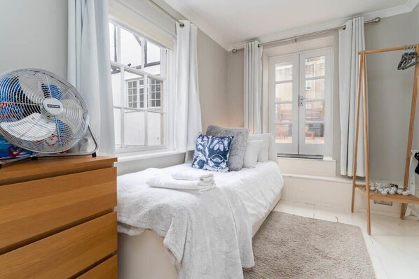 A bedroom with a bed, a wooden dresser, a standing fan, and a window with curtains.