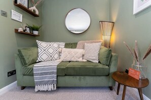 A green sofa with patterned cushions, a round mirror, and a small table with a vase and a welcome sign.