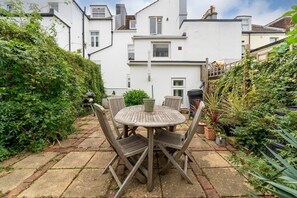 A patio with a round wooden table and chairs, surrounded by potted plants and a hedge.