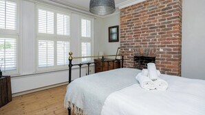 A bedroom with a brick fireplace, a bed with white linens, and a large window with blinds. - Host & Stay | The Walpole House (Brighton and Hove)