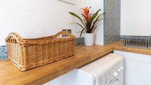 A laundry area with a wicker basket on a wooden counter, a white washing machine, a potted plant, and a framed picture on the wall. - Host & Stay | Islingword Studio (Brighton and Hove)