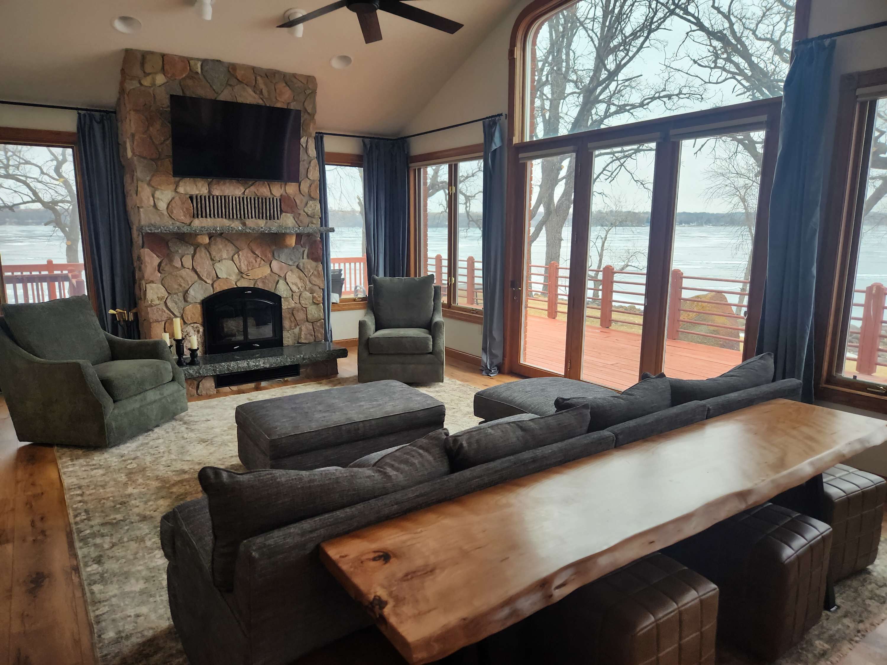 Un salon spacieux doté d’une cheminée en pierre, de sièges confortables et de grandes fenêtres offrant une vue sur un lac.