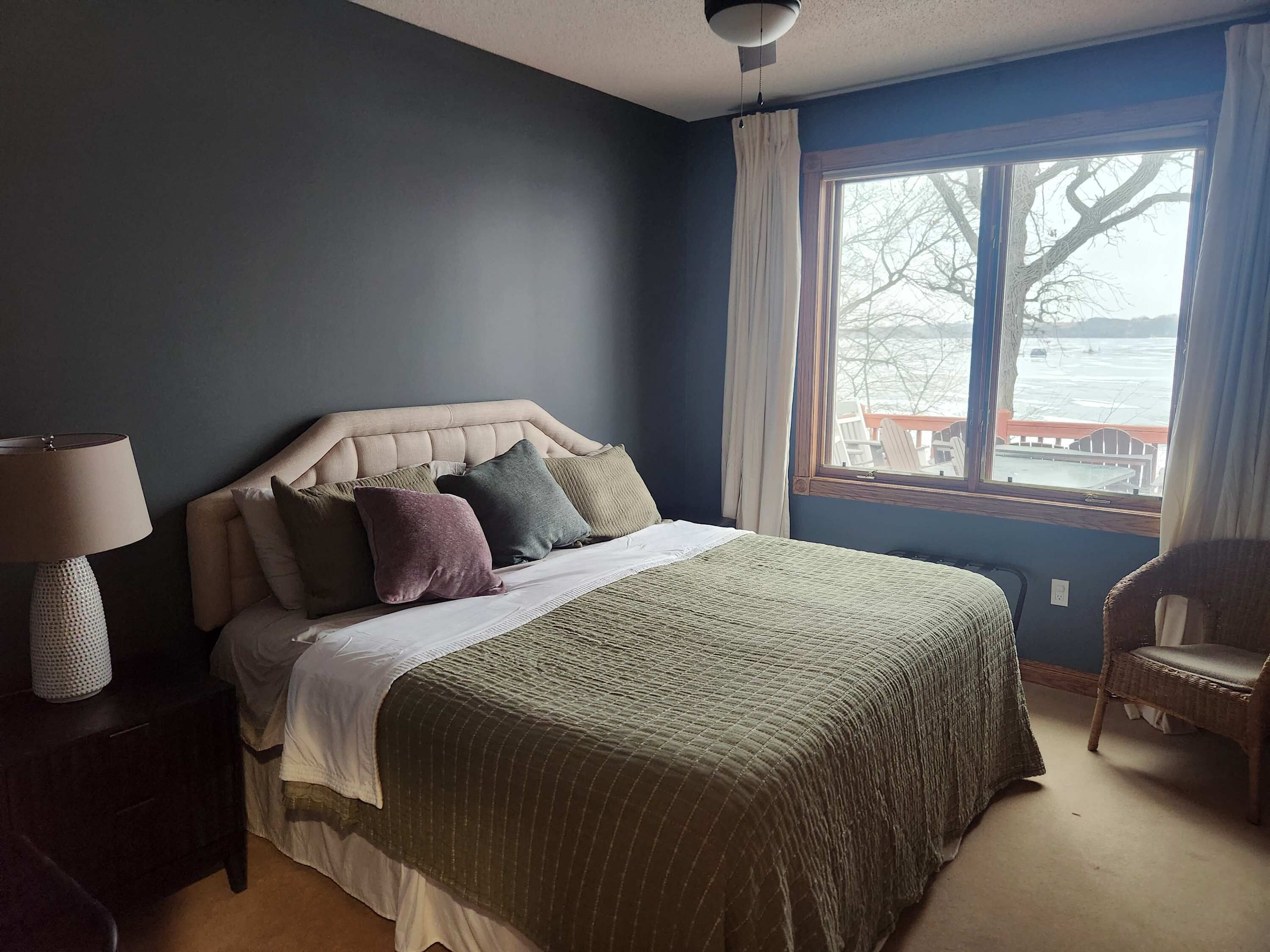 Un dormitorio con cama, una silla, una lámpara y una ventana con vistas a un embarcadero y árboles.