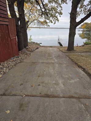 A concrete pathway leading to a body of water, flanked by trees and a red building.