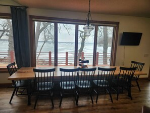 A dining area with a large table and chairs, a hanging light fixture, and a view of a body of water through large windows.